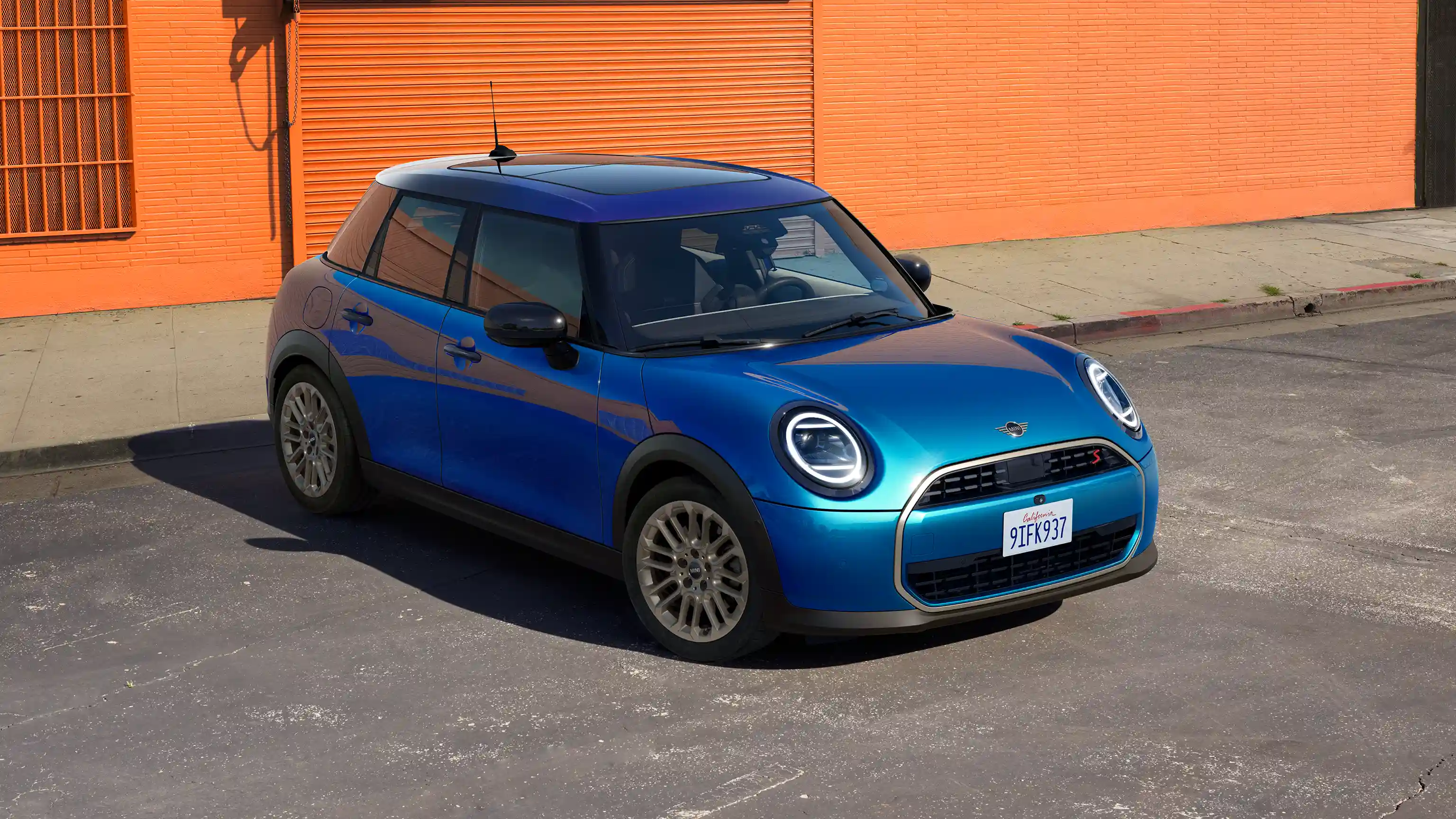Frontansicht eines parkenden MINI Cooper 5-Türer in Icy Sunshine Blue mit Multitone Roof auf einer Straße mit einer orangefarbenen Wand im Hintergrund
