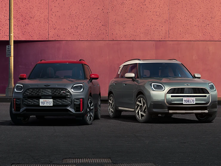 Frontansicht eines MINI Countryman JCW in Legend Grey mit rotem Dach und eines MINI Countryman in Smokey Green mit Dach in Vibrant Silver, die nebeneinander stehen.