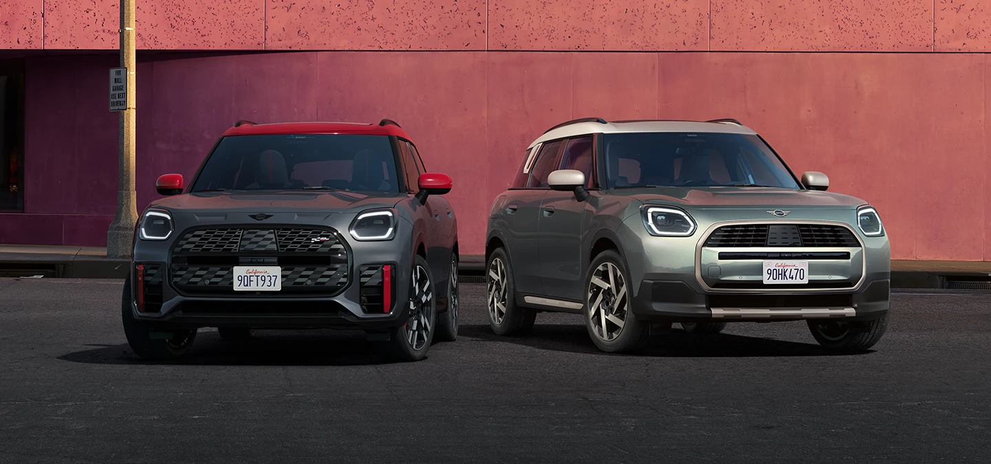 Frontansicht eines MINI Countryman JCW in Legend Grey mit rotem Dach und eines MINI Countryman in Smokey Green mit Dach in Vibrant Silver, die nebeneinander stehen.