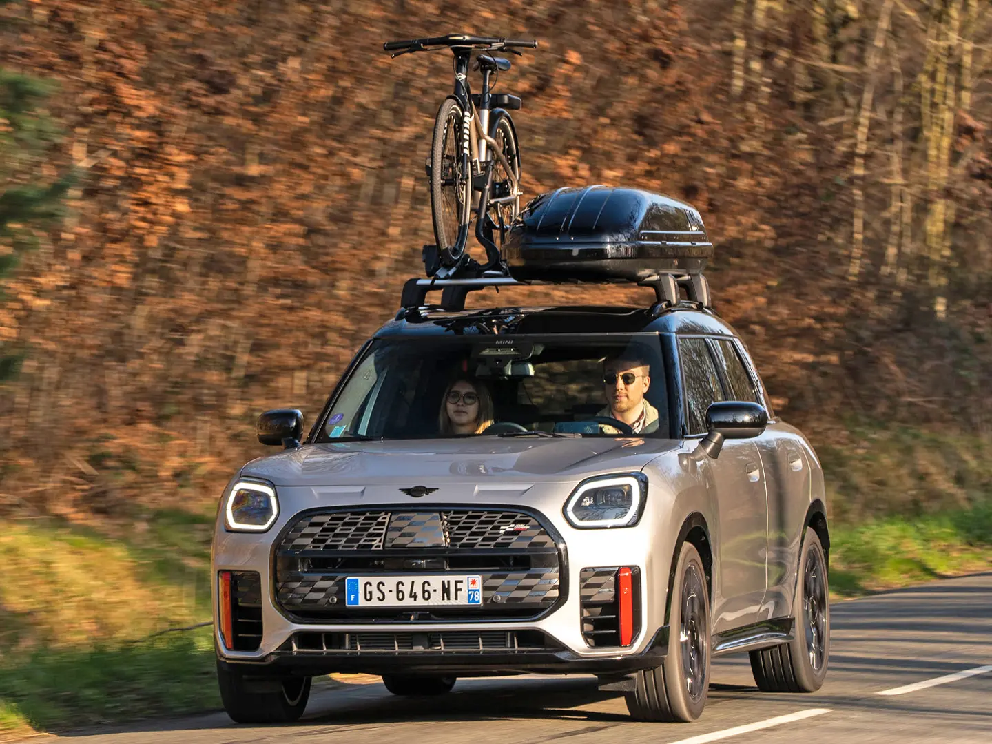 MINI Countryman in Silver auf einer Bergstraße mit MINI Dachträger, MINI Dachbox, MINI Fahrraddachträger mit montiertem Fahrrad und MINI John Cooper Works Winglet in der Frontansicht. 