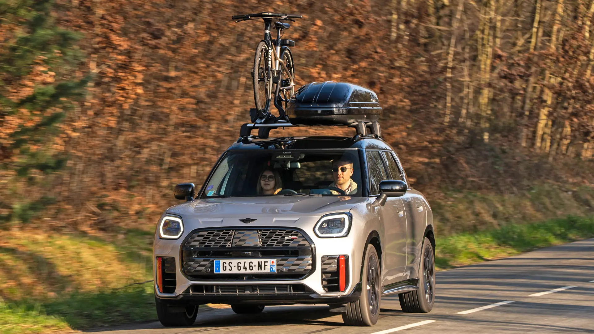 MINI Countryman in Silver auf einer Bergstraße mit MINI Dachträger, MINI Dachbox, MINI Fahrraddachträger mit montiertem Fahrrad und MINI John Cooper Works Winglet in der Frontansicht. 