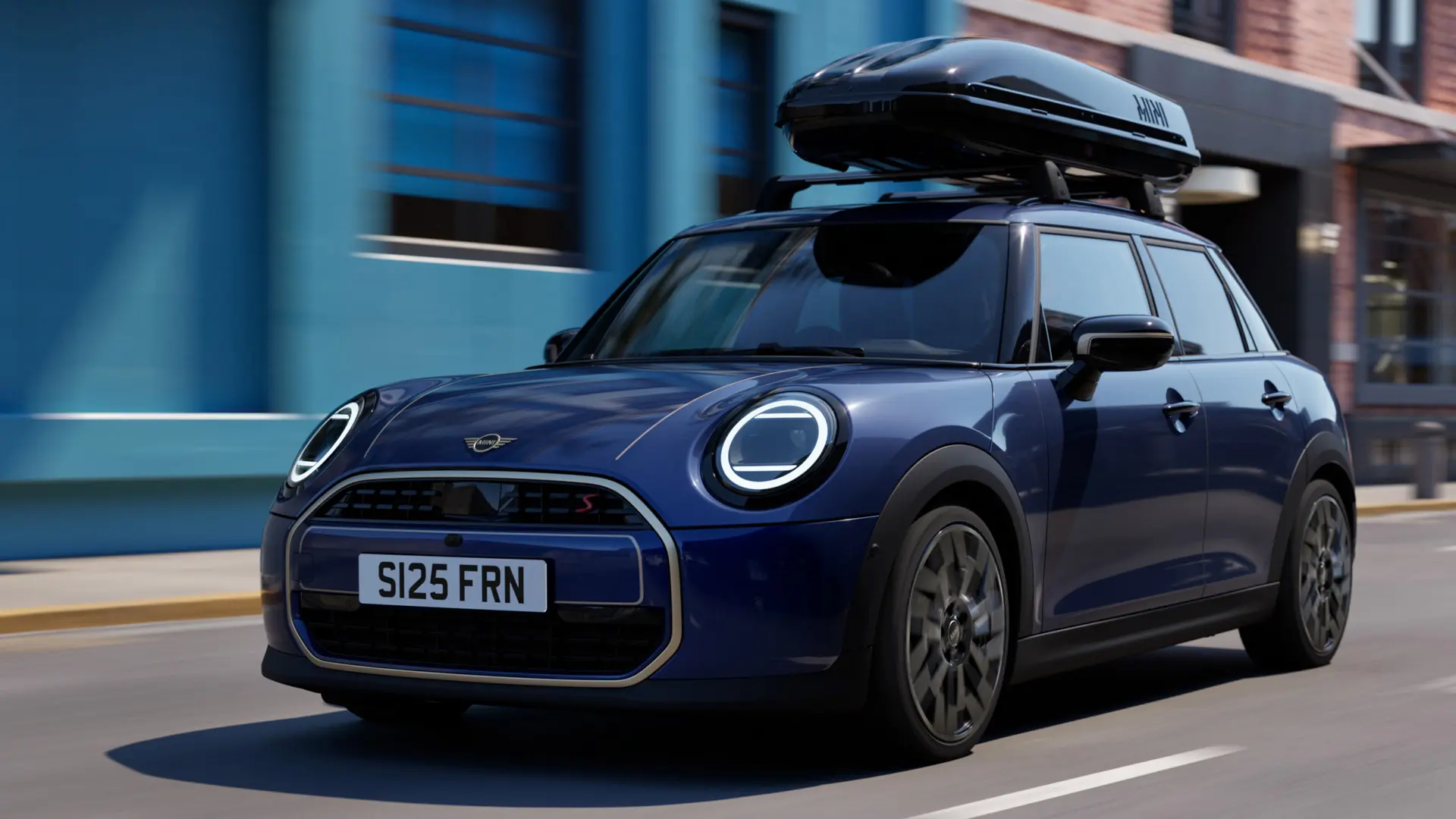 Dreiviertel-Vorderansicht eines MINI Aceman in Indigo Sunset Blue, ausgestattet mit einem MINI Relingträger, auf dem eine MINI Dachbox 320 montiert ist. 