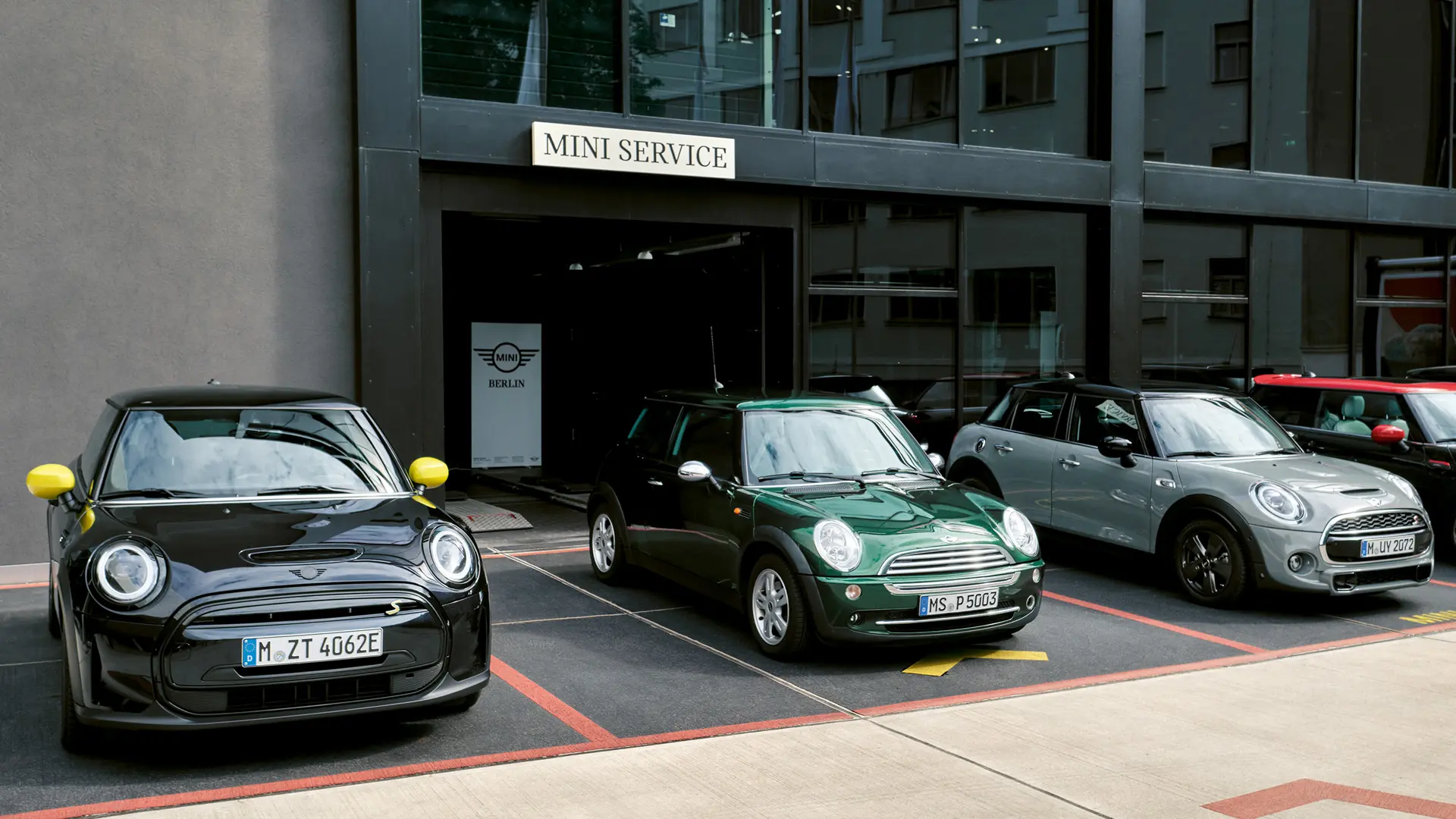 Frontansicht eines MINI Cooper Electric S in Schwarz, der im Freien bei einem MINI Partner neben einem MINI Cooper 3-Türer in Grün, einem MINI 5-Türer in Silber und einem MINI JCW in Schwarz steht. 