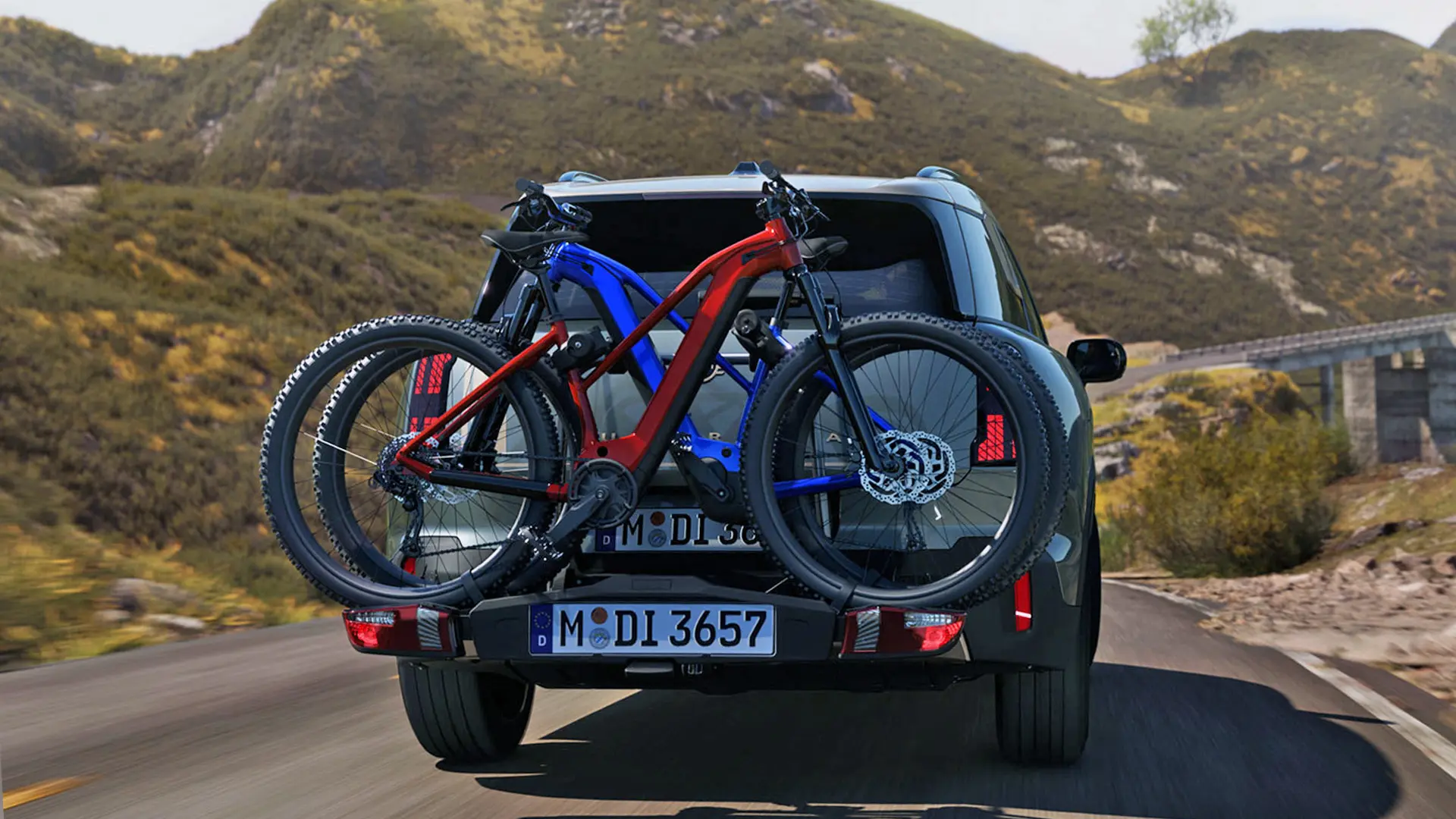 Heckansicht eines MINI Countryman in Smokey Green mit Fahrradheckträger und zwei Mountainbikes, der auf einer Bergstraße fährt.