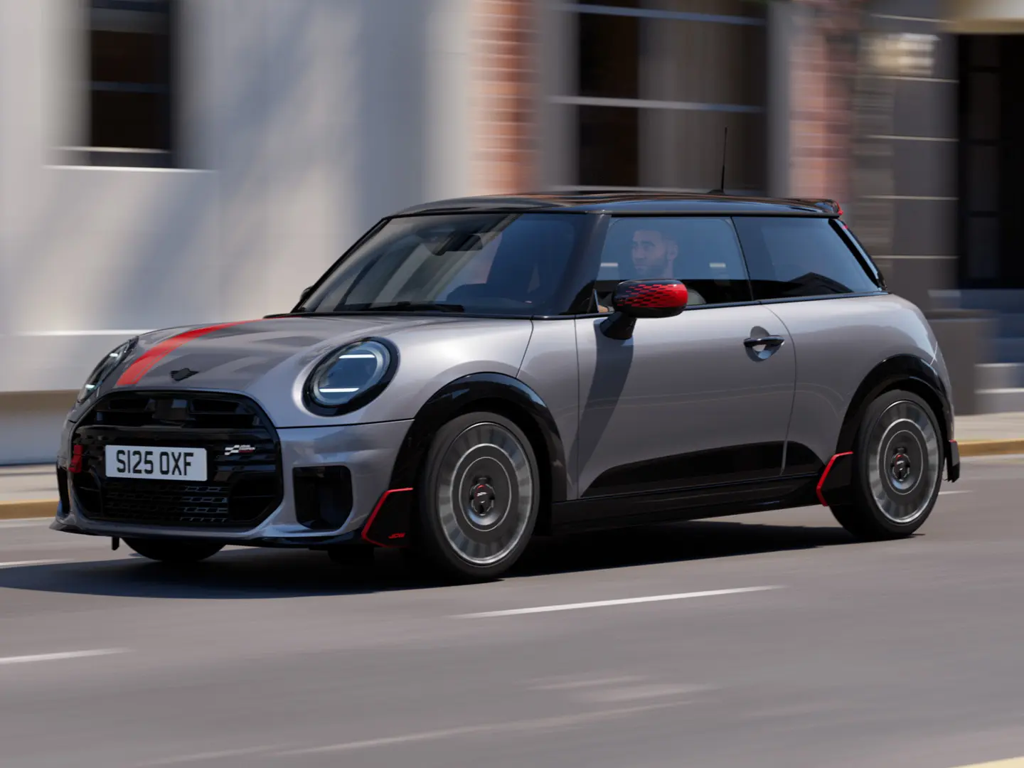 Ein MINI Cooper in Melting Silver mit John Cooper Works Spiegelkappe und Dekoraufklebern auf der Motorhaube in Chili Red, Dachkantenspoiler und Winglet in Schwarz fährt auf einer Straße in der Stadt – seitliche Frontansicht.