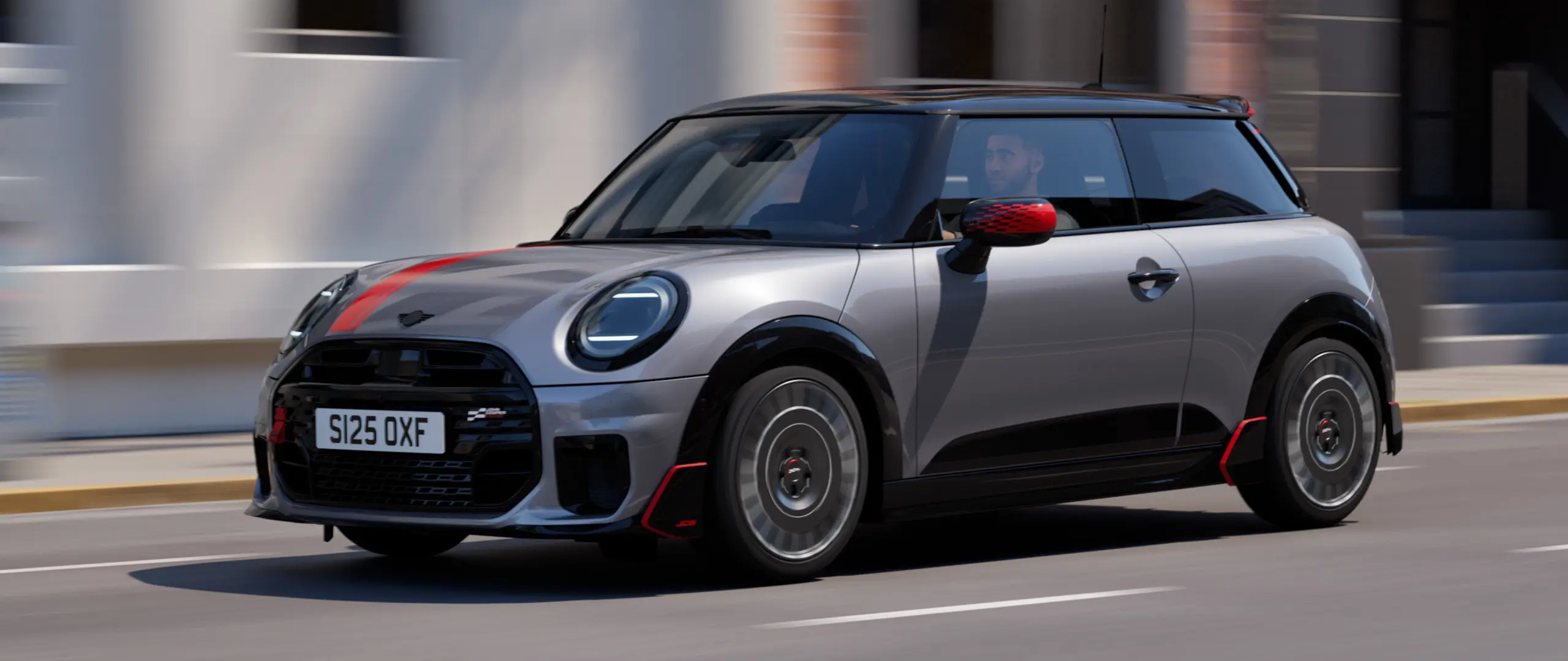 Ein MINI Cooper in Melting Silver mit John Cooper Works Spiegelkappe und Dekoraufklebern auf der Motorhaube in Chili Red, Dachkantenspoiler und Winglet in Schwarz fährt auf einer Straße in der Stadt – seitliche Frontansicht.