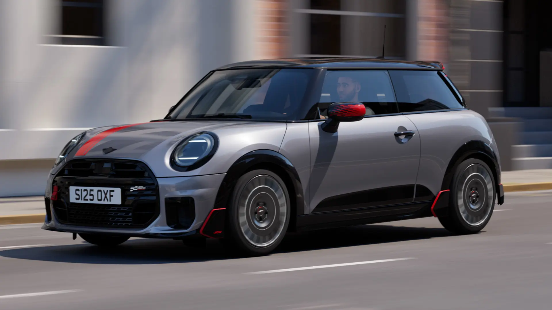 Ein MINI Cooper in Melting Silver mit John Cooper Works Spiegelkappe und Dekoraufklebern auf der Motorhaube in Chili Red, Dachkantenspoiler und Winglet in Schwarz fährt auf einer Straße in der Stadt – seitliche Frontansicht.