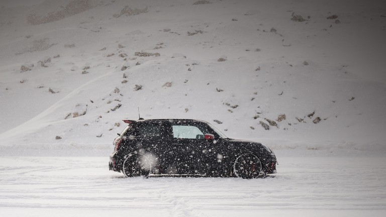 Eine Person geht über eine verschneite Fläche auf einen schwarzen MINI Cooper zu, der vor einer winterlichen Berglandschaft steht.