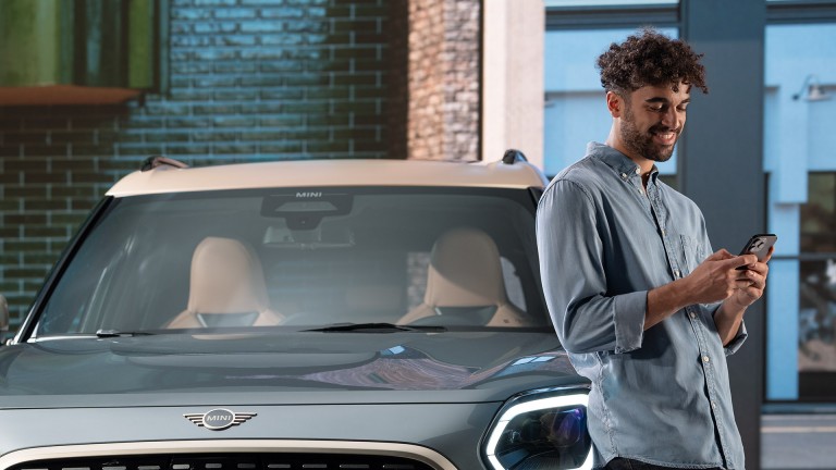 Ein junger Mann lehnt sich lächelnd an seinen MINI Countryman und sieht dabei in sein Handy