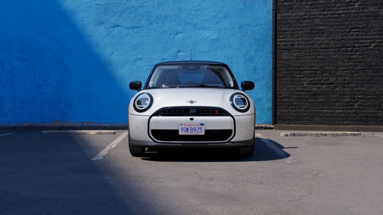 Ein weißer MINI Cooper steht frontal auf einem Parkplatz vor einer blauen Wand.